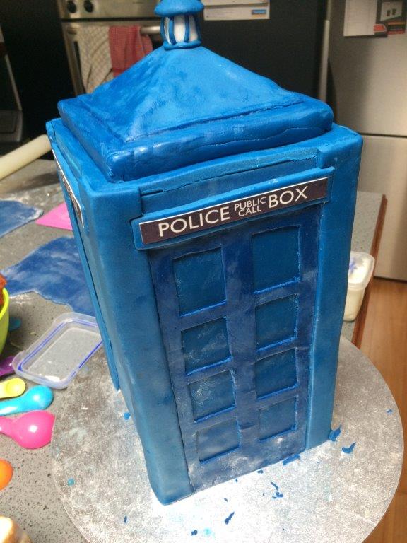Dr Who Tardis Cake | The TARDIS is Homemade fondant on an “orange velvet” cake filled and covered in dark chocolate orange ganache. The board is covered in white fondant then baked for 10 minutes at 80C to set it hard before it was airbrushed with food colour to look like a galaxy. The signage is icing sheets printed with edible ink. I made the Dalek from homemade modelling chocolate. | https://robertscakesandcooking.com/tardis-cake-and-construction-photos/